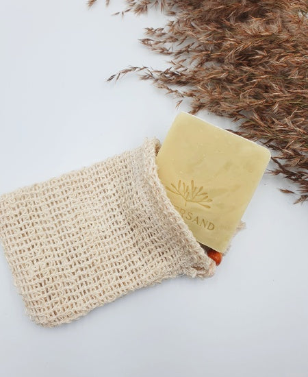 Eine helle Naturseife ragt aus einem Sisal-Säckchen auf weißem Hintergrund mit Schilfgras
